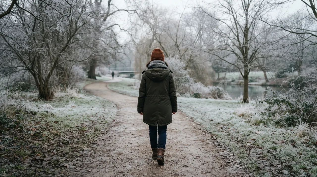 Person in Winterjacke und Mütze geht an einem bedeckten Wintertag einen Parkweg mit Raureif entlang